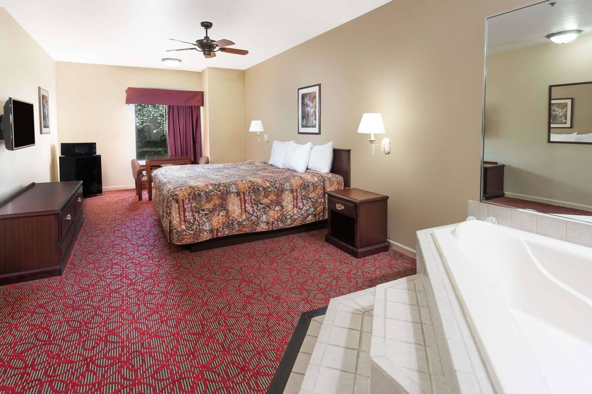 A hotel room with a bed, a desk, a chair, a TV, a ceiling fan, a window with curtains, a bathroom with a bathtub, and a mirror.
