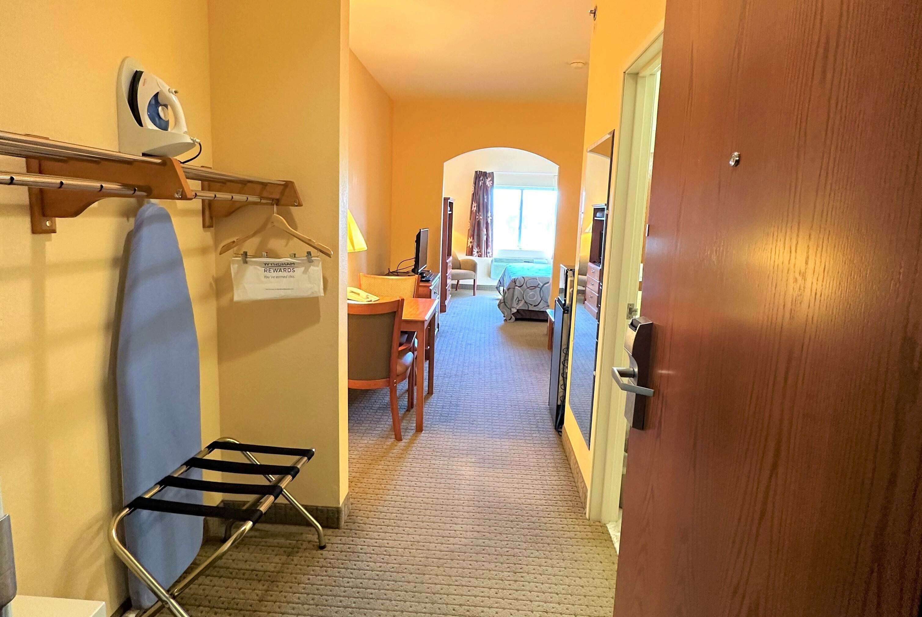 A hotel corridor with a wooden door, a hanging iron, a towel rack, a chair, a desk with a lamp, and a bed in the background.