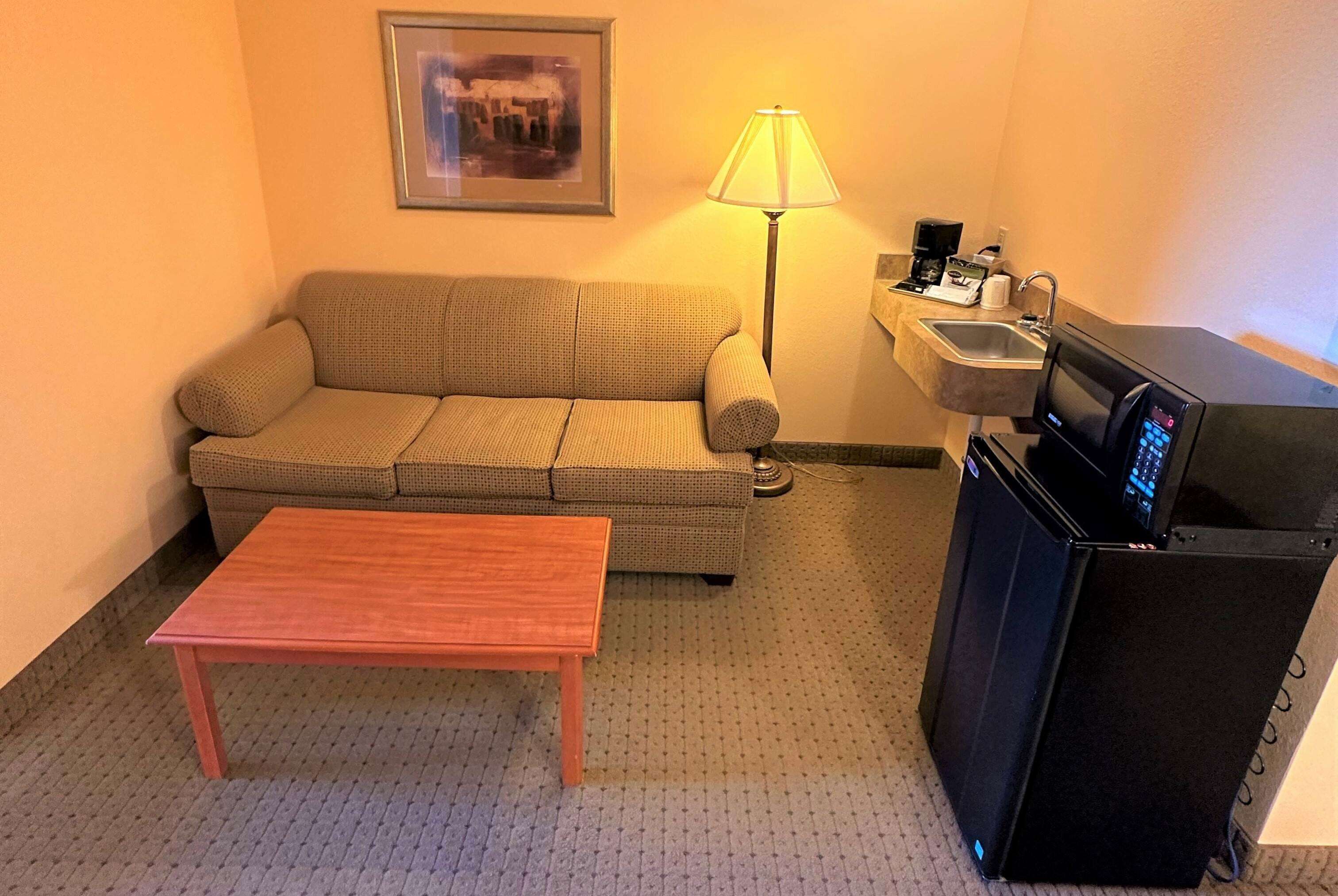 Una habitación de hotel pequeña con sofá, una mesa de madera, lámpara, lavamanos y microondas.
