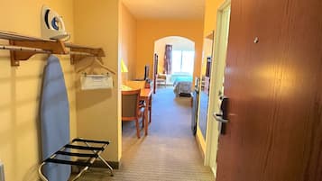 A hotel corridor with a wooden door, a wall-mounted ironing board, a hanging towel rack, a small table with chairs, and a view into a room with a bed and window.