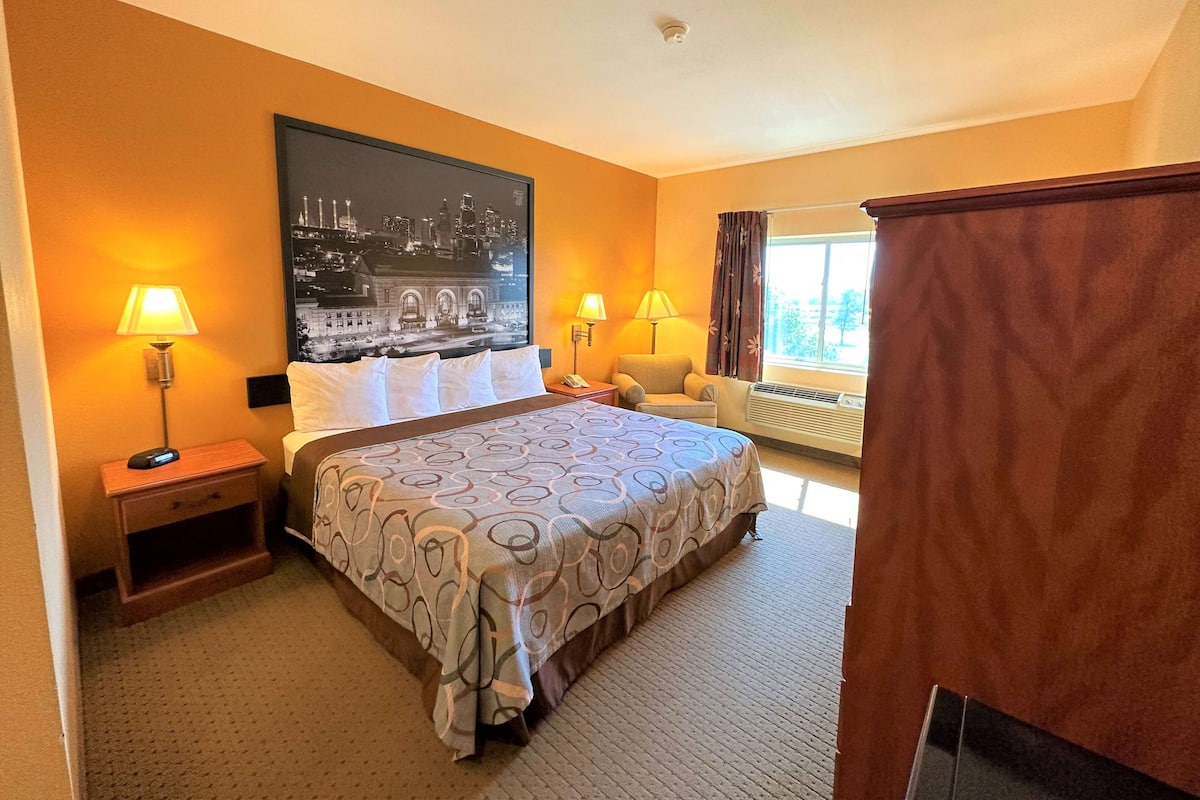 Ein Hotelzimmer mit einem großen Bett, zwei Nachttischlampen, einem Sessel, einem Fenster mit Vorhängen und einem Stadtbild an der Wand.