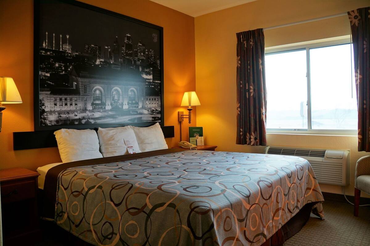 Ein Hotelzimmer mit einem großen Bett, zwei Nachttischlampen, einem Nachttisch, einem Fenster mit Vorhängen und einem Stadtplakat an der Wand.