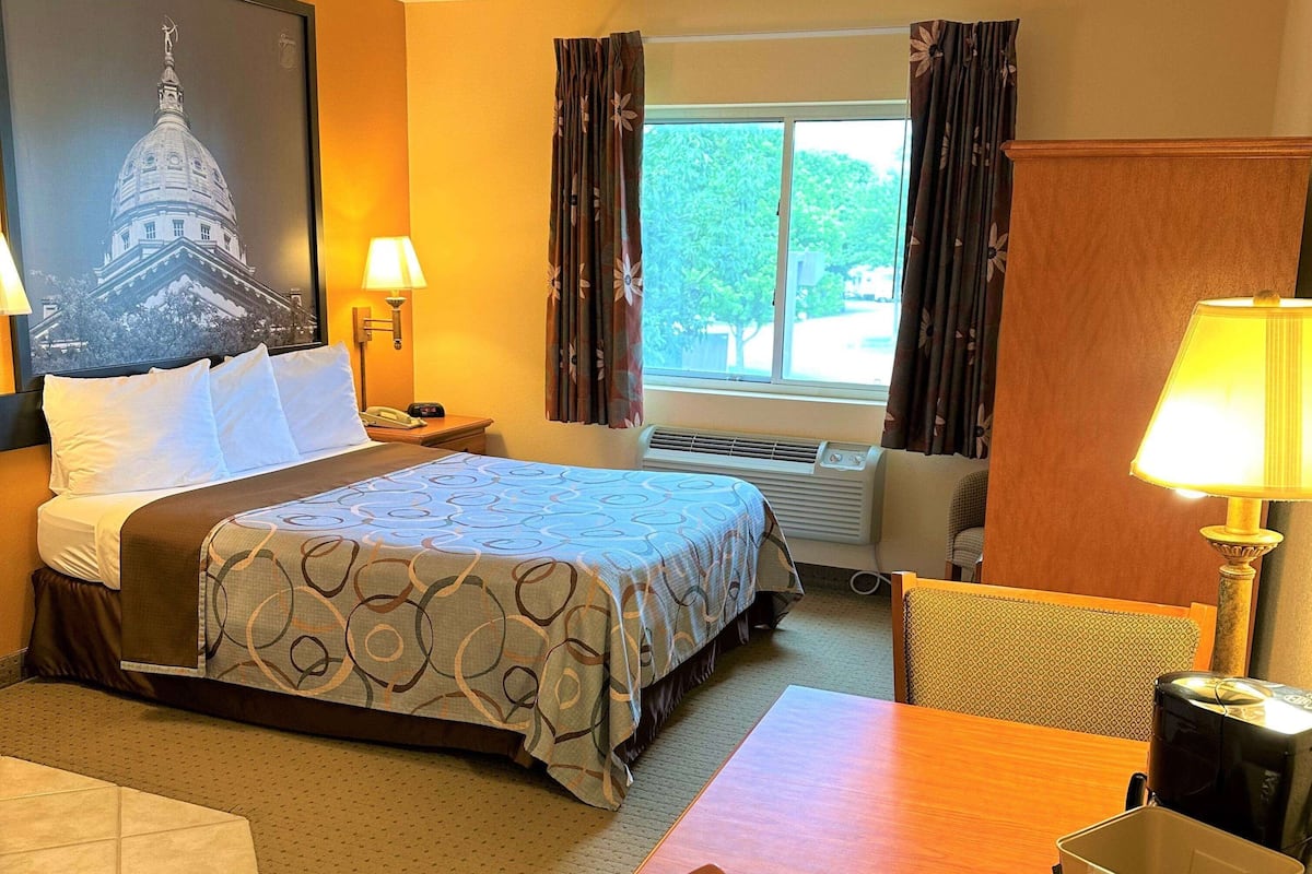 Ein Hotelzimmer mit Bett, Schreibtisch, Stuhl, einem Fenster mit Vorhängen, einer Wandlampe und einem Gemälde eines Gebäudes.