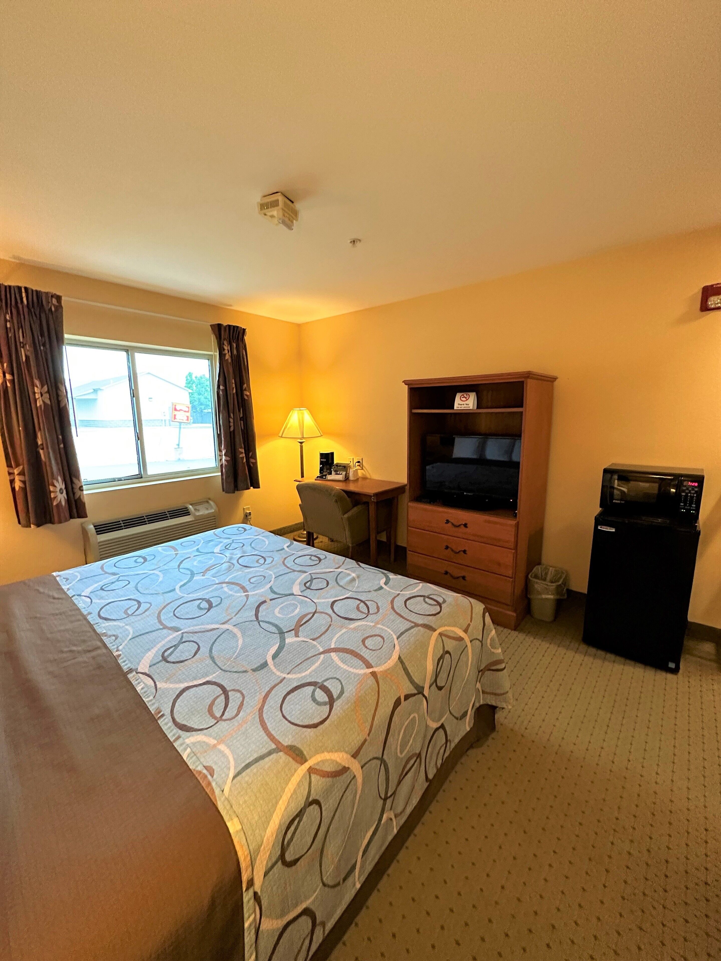A hotel room with a bed, a desk, a TV, a refrigerator, and a window with curtains.