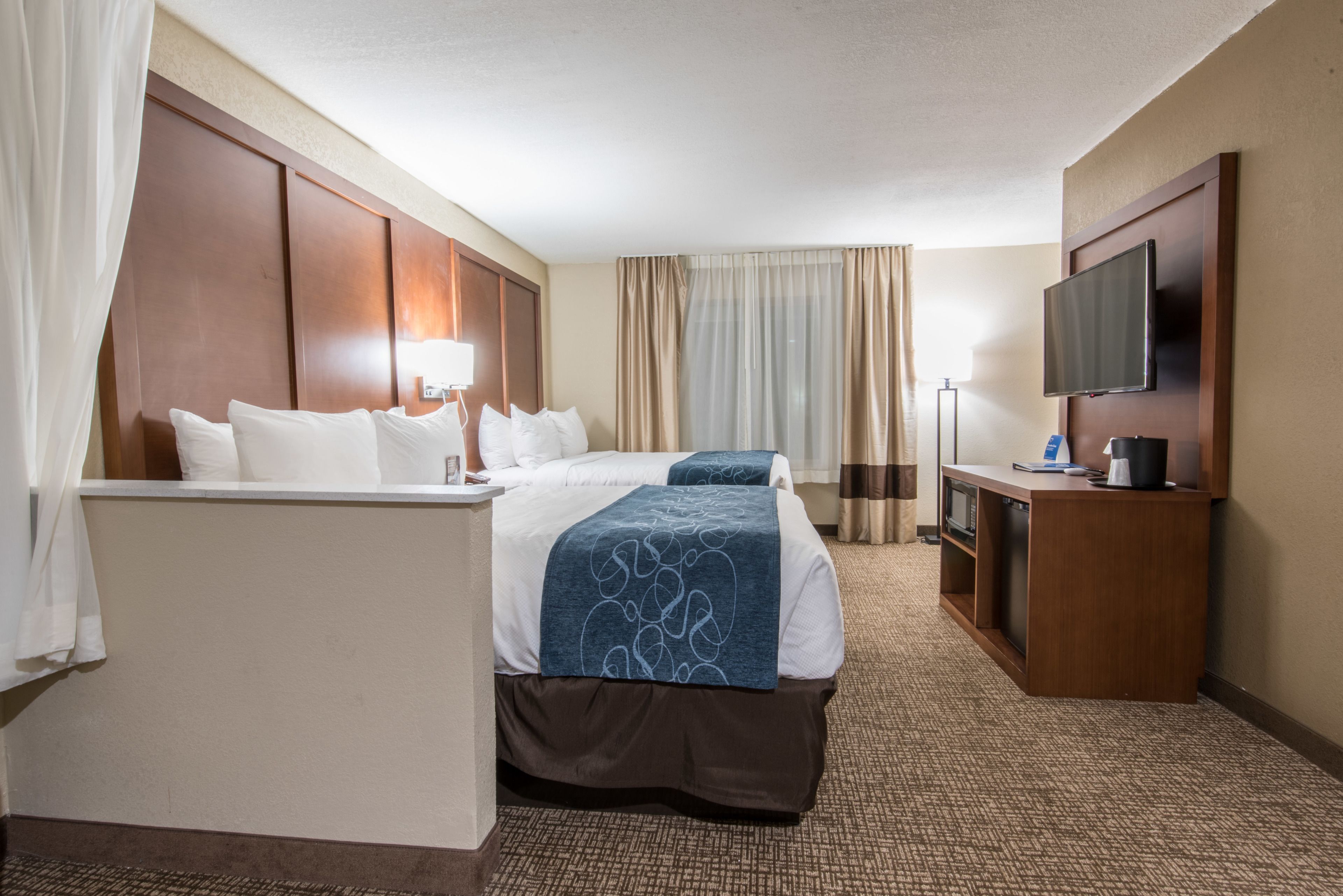 Ein Hotelzimmer mit zwei Betten, einem Schreibtisch, einem Fernseher und einem großen Fenster mit Vorhängen.