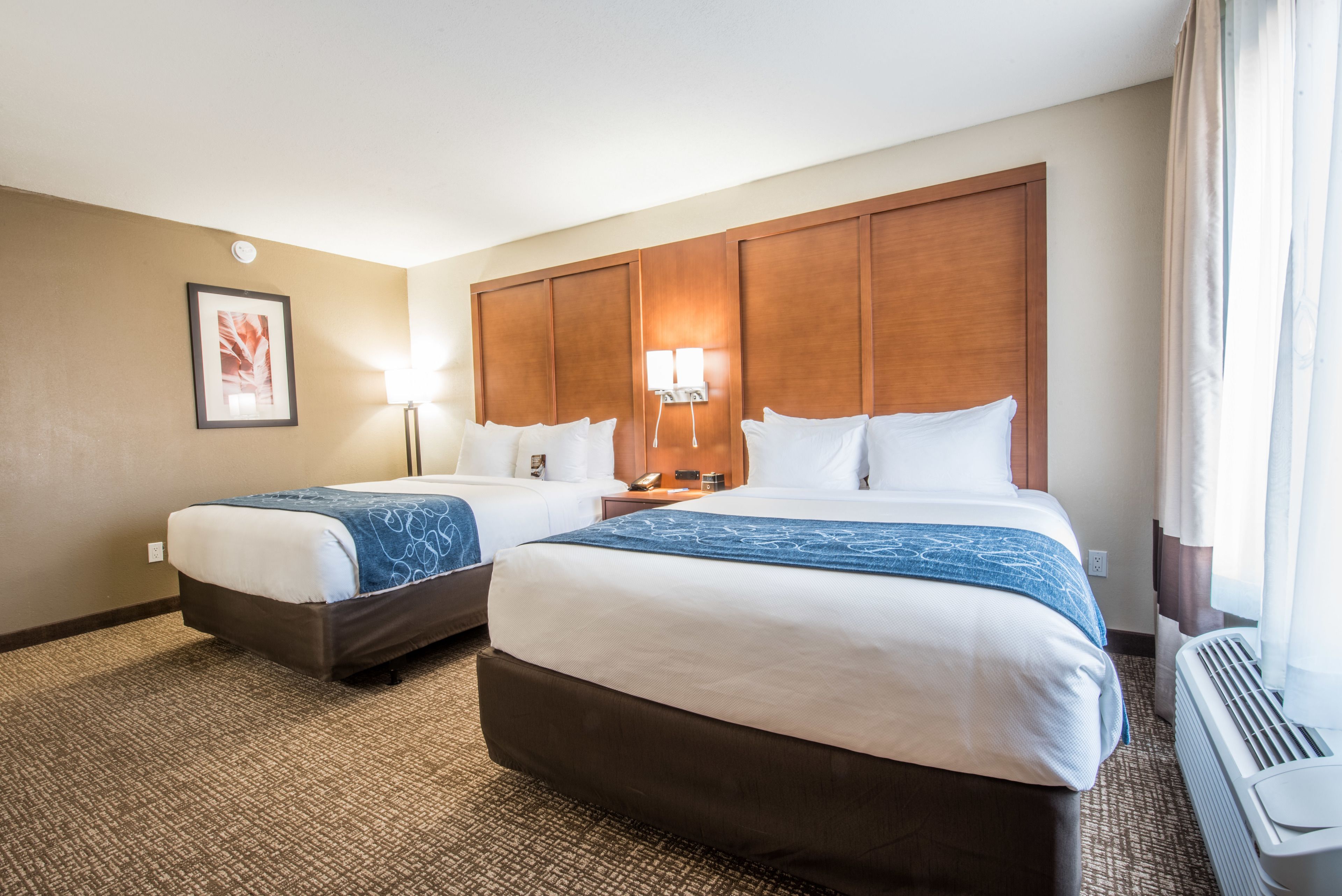 A hotel room with two beds, a large wooden headboard, a framed picture on the wall, a floor lamp, and a window with curtains.