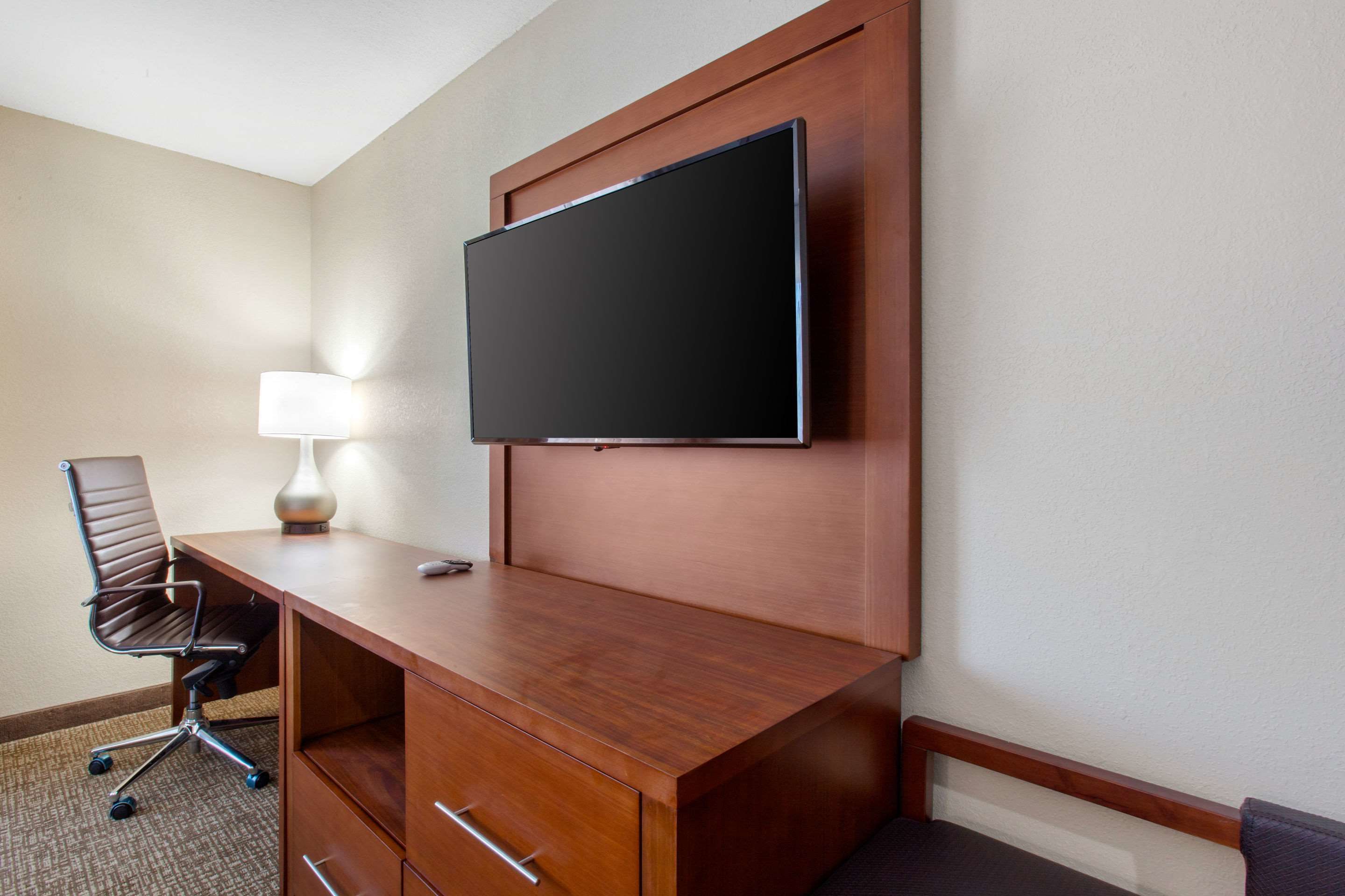A hotel room with a wooden desk, a flat-screen TV mounted on the wall, a modern chair, and a desk lamp.