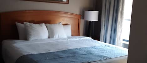 Habitación de hotel con una cama grande, cabecera, un cuadro en la pared, una lámpara y una ventana con cortinas.