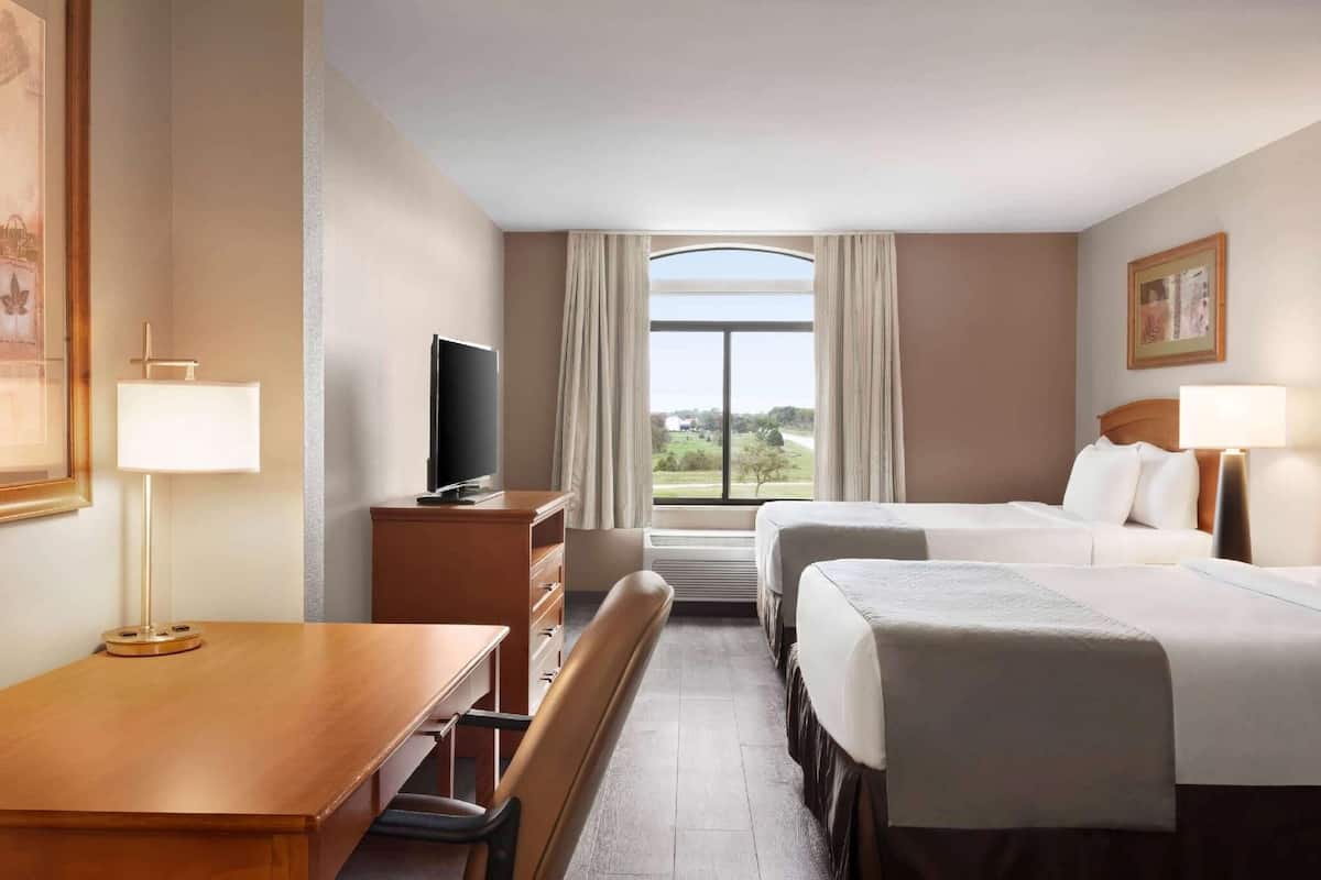 A hotel room with a desk, two beds, a TV, a window with curtains, and a painting on the wall.