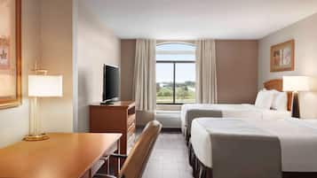 Quarto de hotel com duas camas, uma escrivaninha com cadeira, televisão e janela com cortinas.
