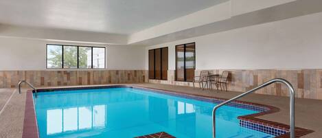 An indoor swimming pool with clear blue water, a tiled border, and a seating area with chairs and a table.