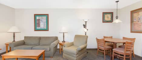 A living room with a sofa, armchair, dining table, and chairs. There are lamps and framed pictures on the walls.