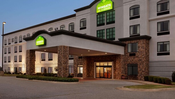 A Wingate by Wyndham hotel with a stone facade, multiple windows, and a well-lit entrance. - Wingate by Wyndham Bentonville AR (Bentonville)