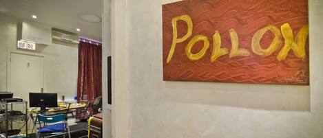 A room with a large painting of the word "POLLON" on the wall, a desk with a computer, a chair, and a small table with a lamp.