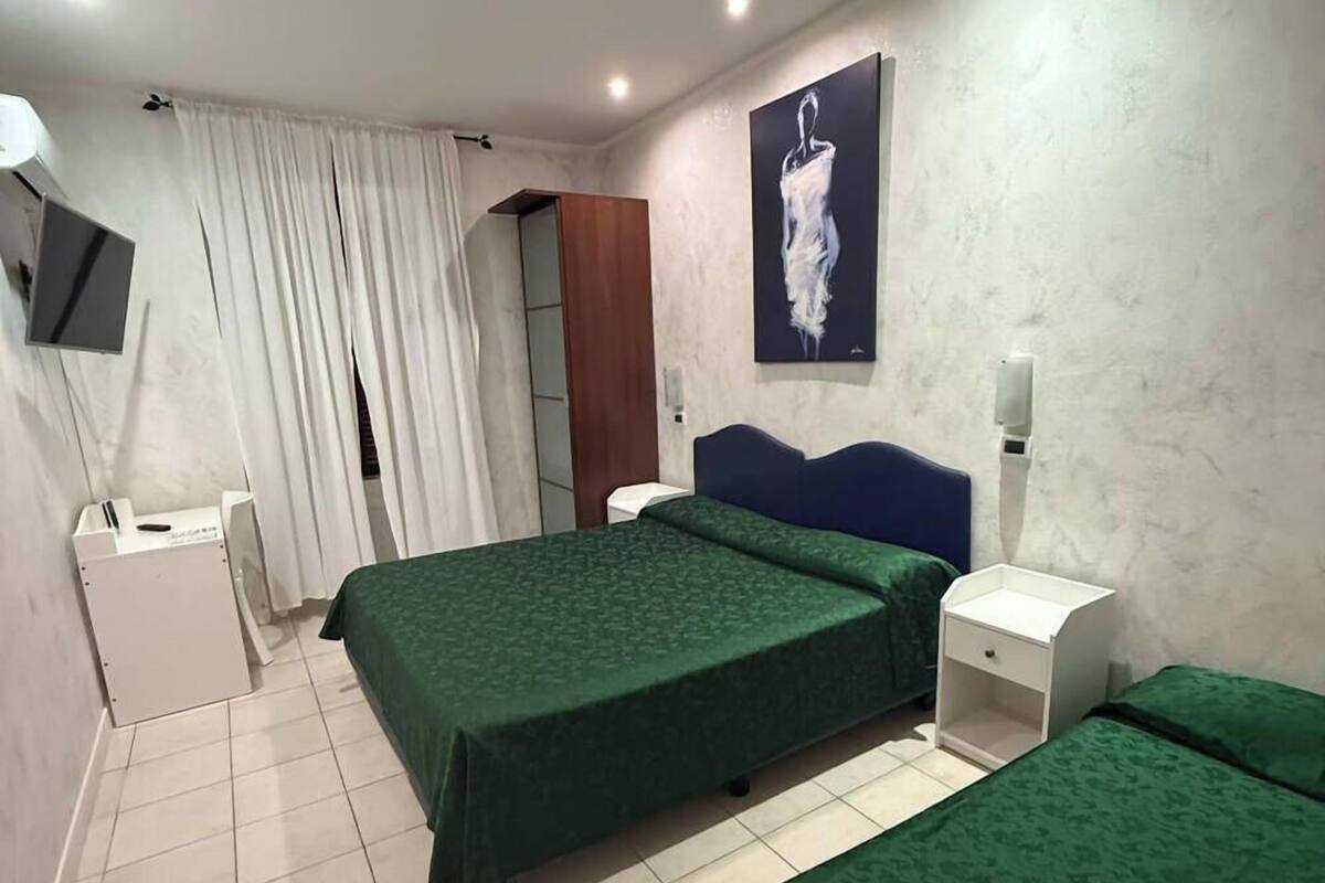 Habitación de hotel con dos camas individuales, un televisor montado en la pared, un armario y una mesita de noche con lámpara.