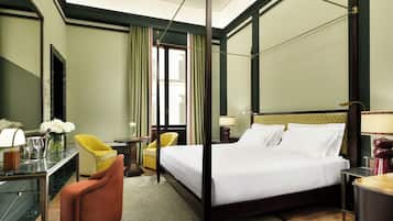 Quarto de hotel com cama de dossel, escrivaninha com cadeira, mesinha de canto com abajur e poltrona com um vaso de flores.