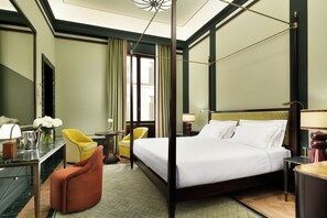 A hotel room with a four-poster bed, a desk with a chair, a small table with a lamp, and a chair with a vase of flowers. - Palazzo Ruspoli (Florence)