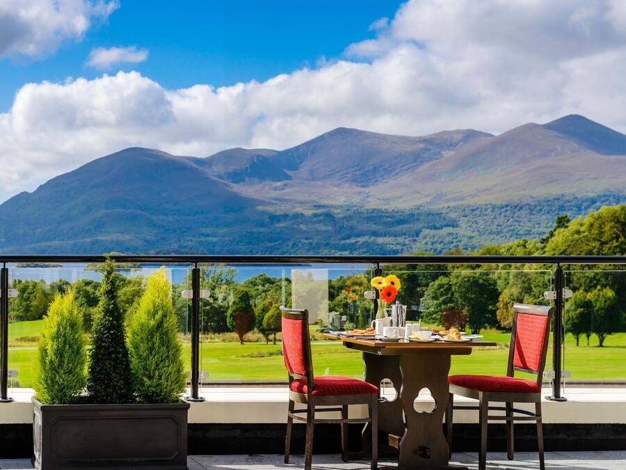 A balcony with a table and chairs, overlooking a scenic landscape with mountains and a lake.