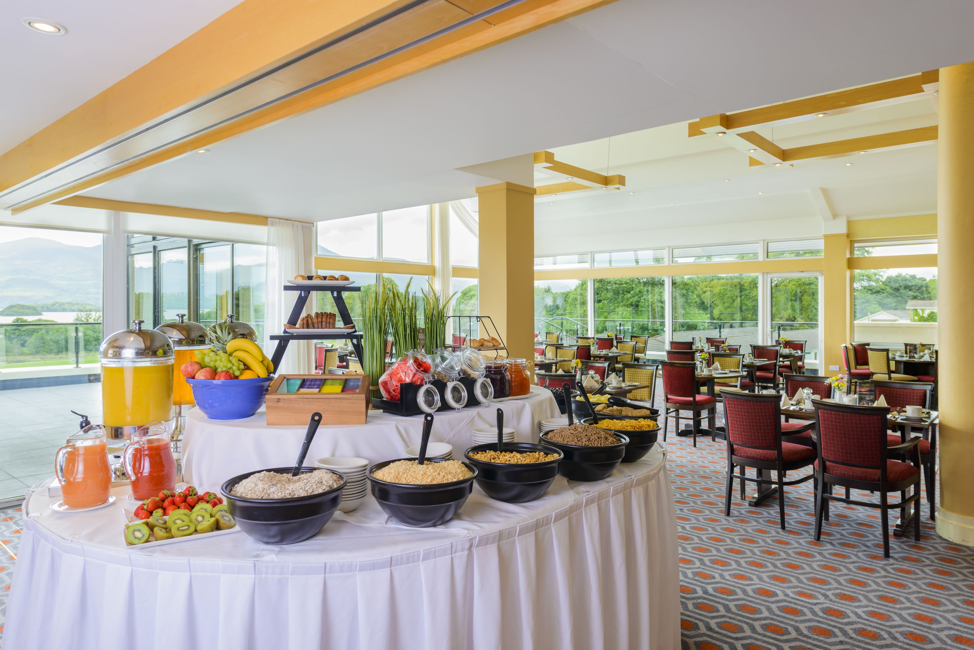 A buffet setup with various food items and beverages, a large window with a view of greenery, and a spacious dining area with chairs.