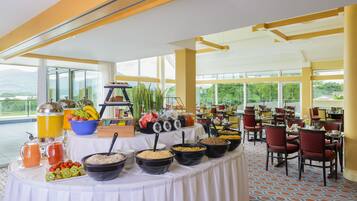 A buffet setup with various food items and beverages, a large window with a view of greenery, and a spacious dining area with chairs.