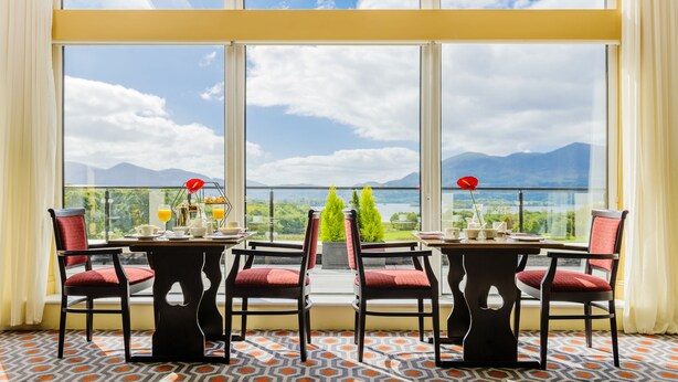 A dining area with a table set for two, chairs, and a view of mountains through large windows. - Castlerosse Park Resort (Killarney)