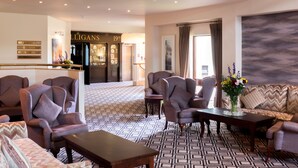 A hotel lobby with a patterned floor, multiple armchairs, a sofa, a coffee table, and a reception area with a sign reading "ALLIGANS 19th". - Castlerosse Park Resort (Killarney)