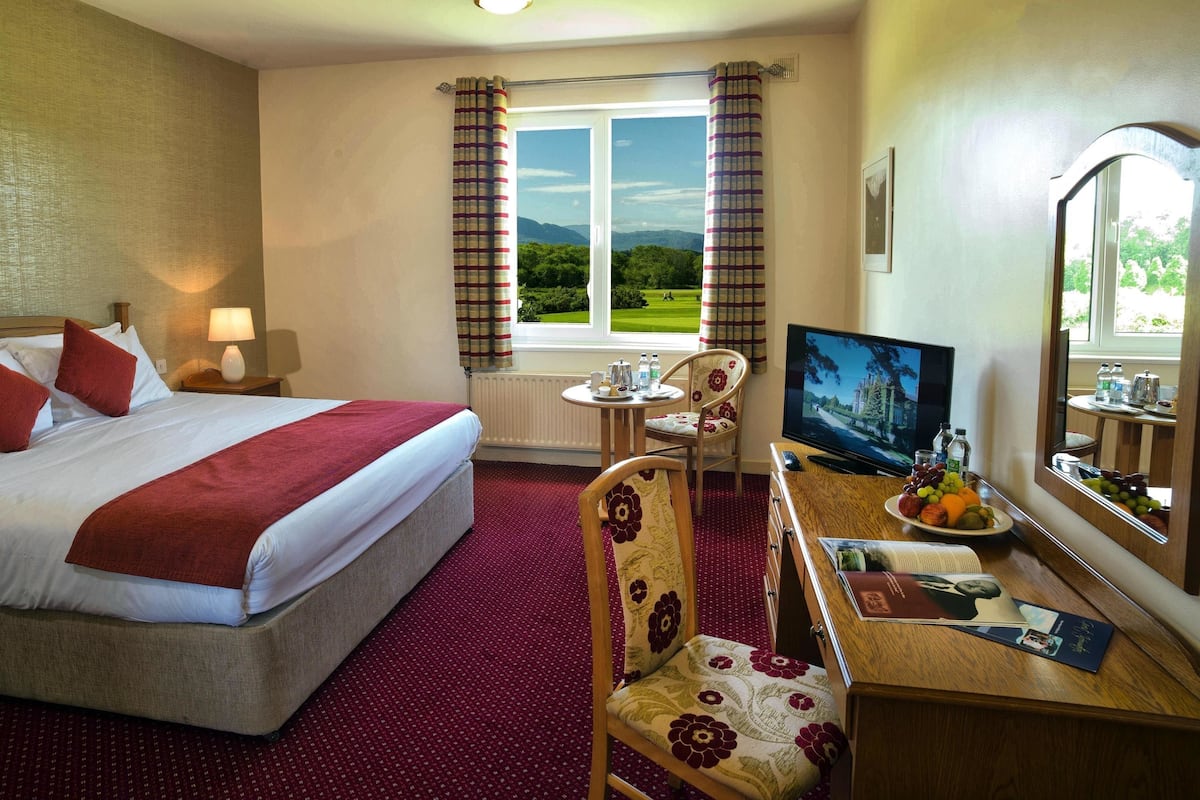 Habitación de hotel con cama, escritorio con computadora, televisión, silla y ventana con cortinas.