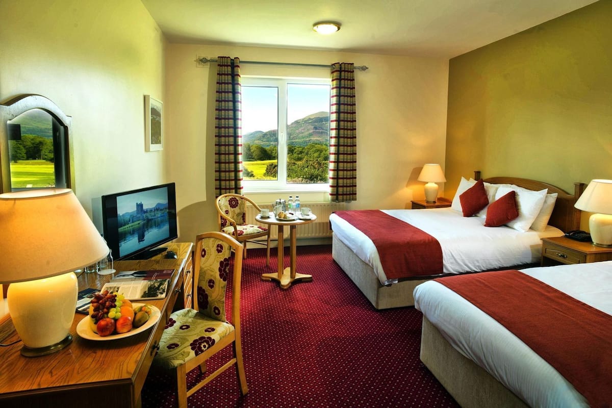 Una habitación de hotel con dos camas, un escritorio con ordenador, una silla, una mesa con un plato de fruta y vistas a un paisaje montañoso.