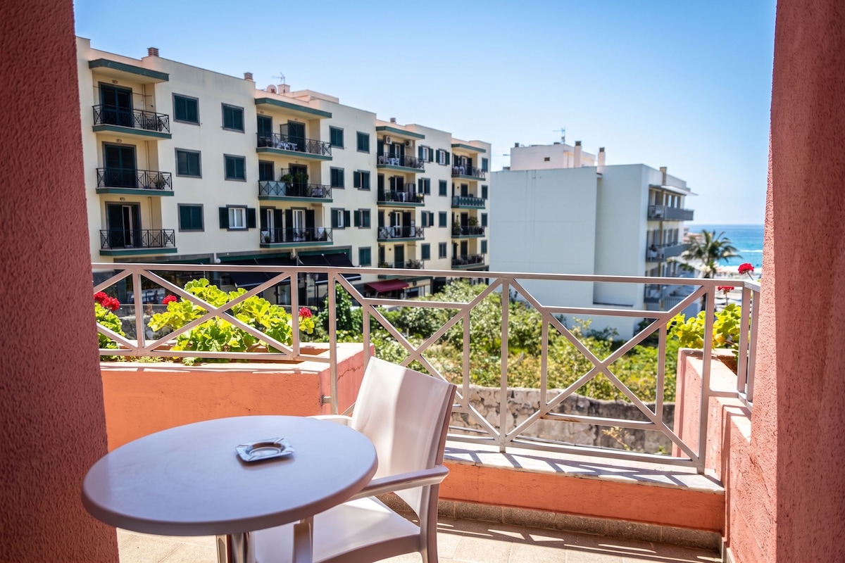 A balcony with a table and chair, overlooking a coastal area with buildings and a clear sky.