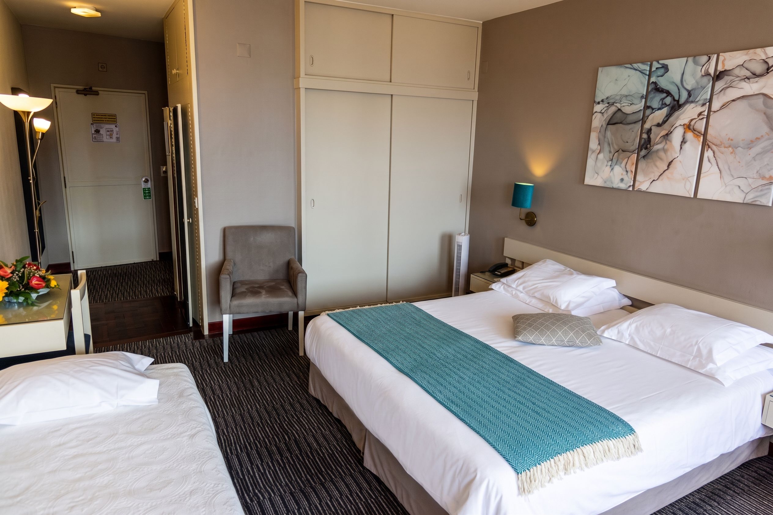 A hotel room with a bed, a chair, a desk with a lamp, and a painting on the wall.