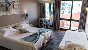 A hotel room with two beds, a small table with chairs, a large window with curtains, and a view of buildings outside.