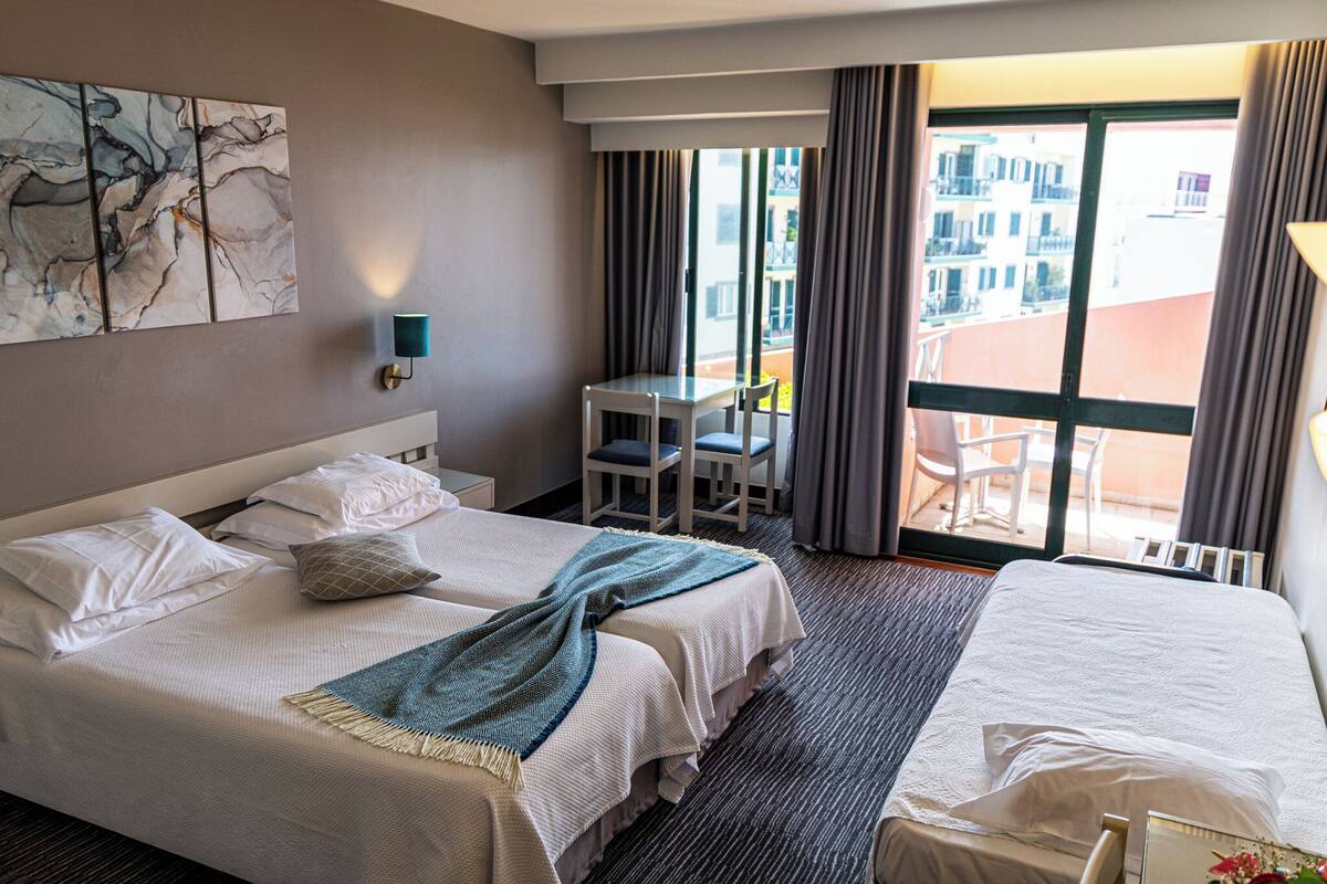 A hotel room with two beds, a small table with chairs, a large window with curtains, and a view of buildings outside.