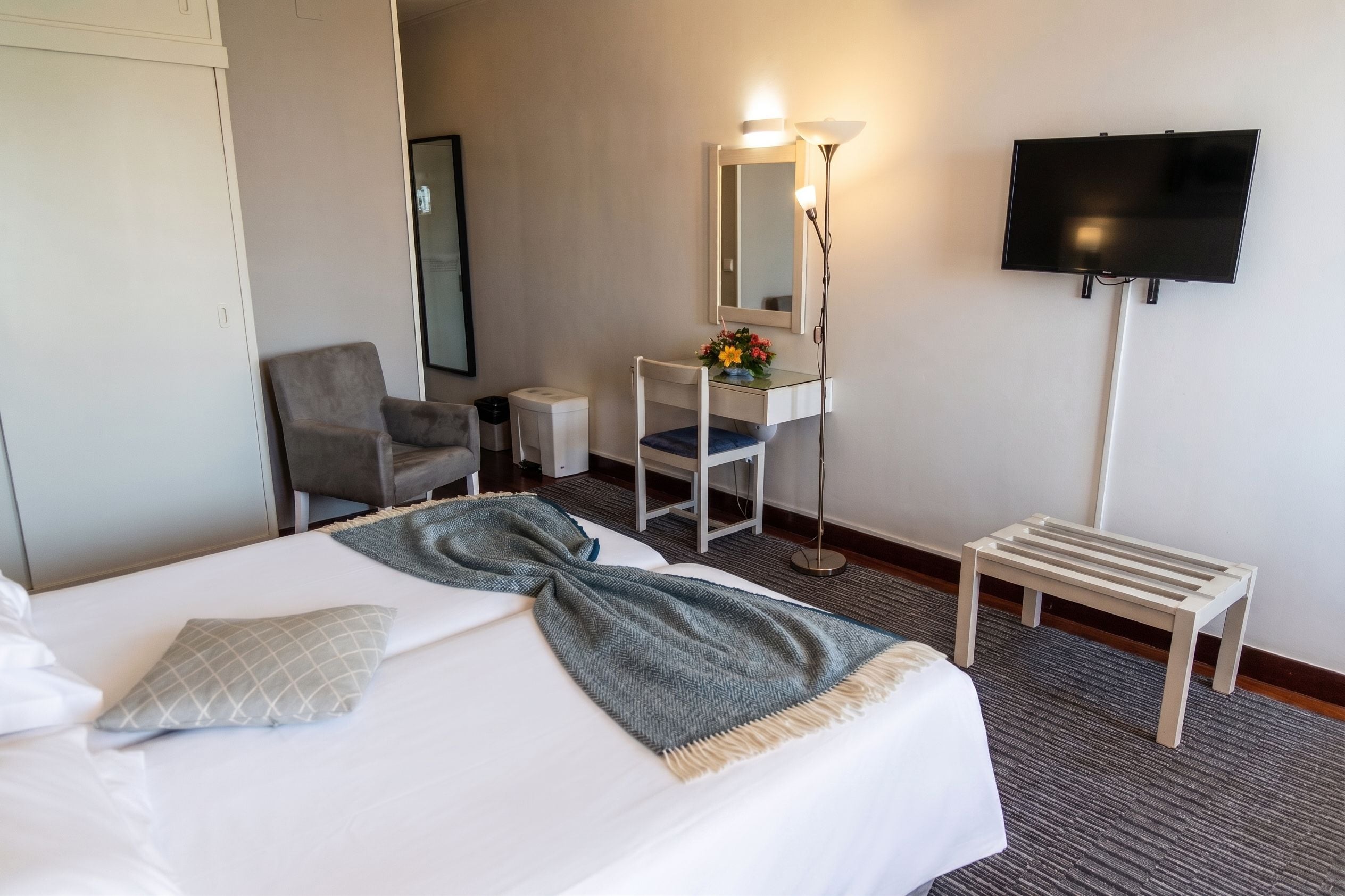 A hotel room with a bed, a desk with a mirror, a chair, a TV, and a small table.