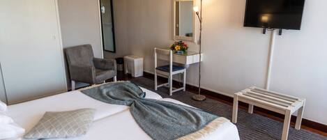 A hotel room with a bed, a desk with a mirror, a chair, a TV, and a small table.