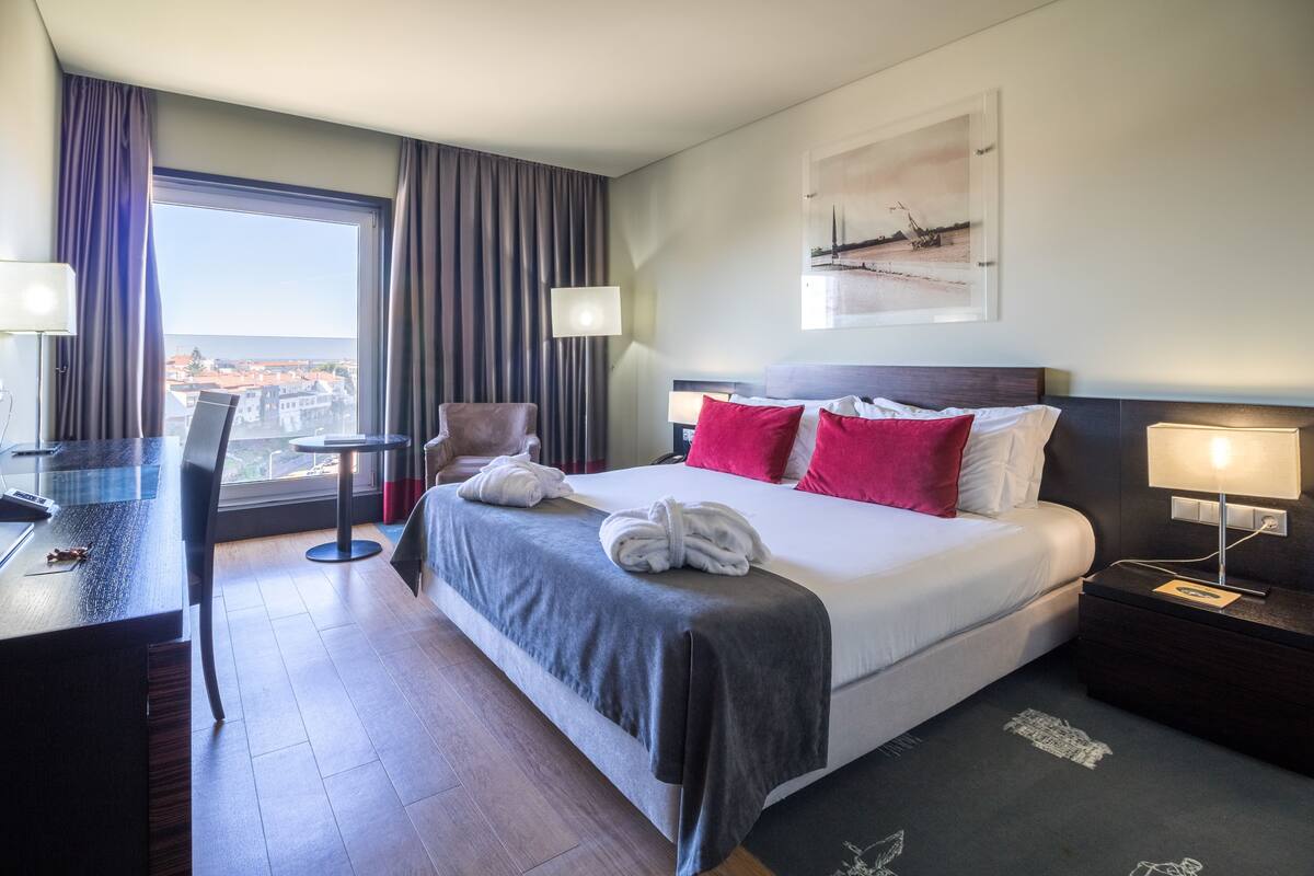 A hotel room with a large bed, a desk, a chair, and a view of the city through the window.