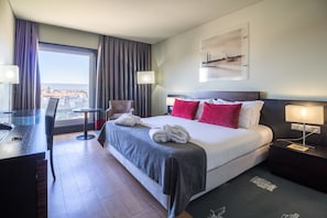 A hotel room with a large bed, a desk, a chair, and a view of the city through the window.