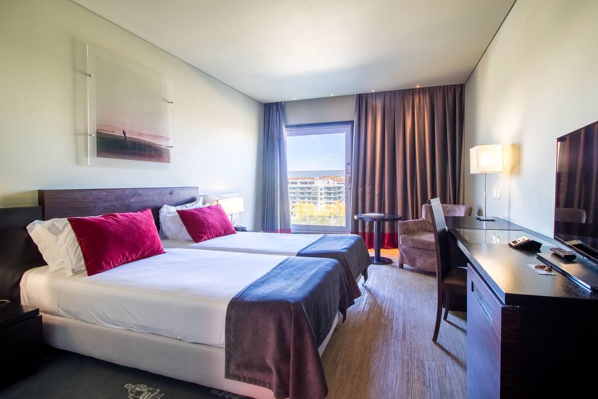 A hotel room with two beds, a desk, a chair, a TV, and a window with curtains.
