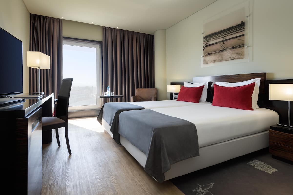 A hotel room with a large bed, two bedside tables, a desk, a chair, and a window with curtains.