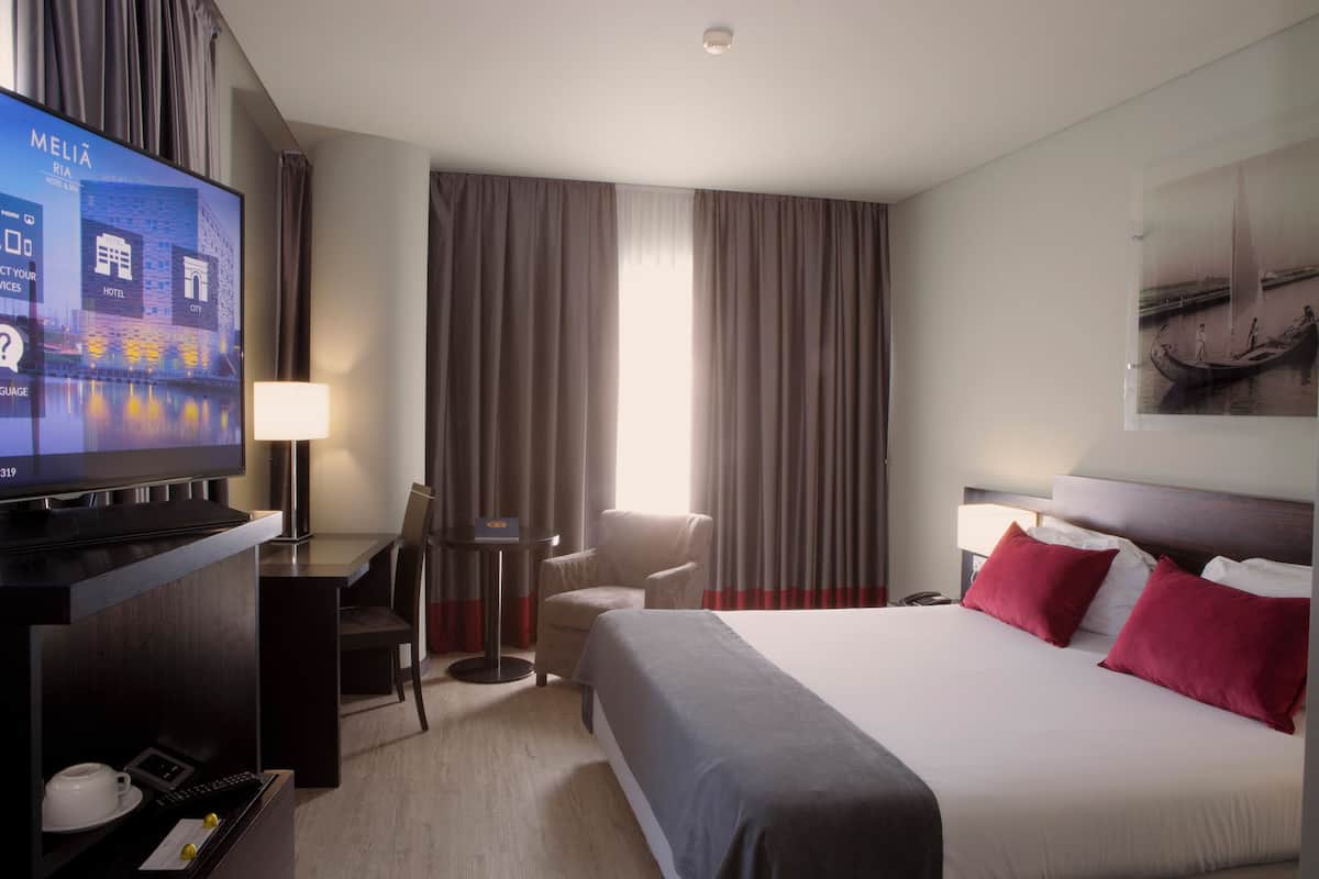 A hotel room with a bed, a desk with a computer, a TV, a chair, a lamp, and a window with curtains.
