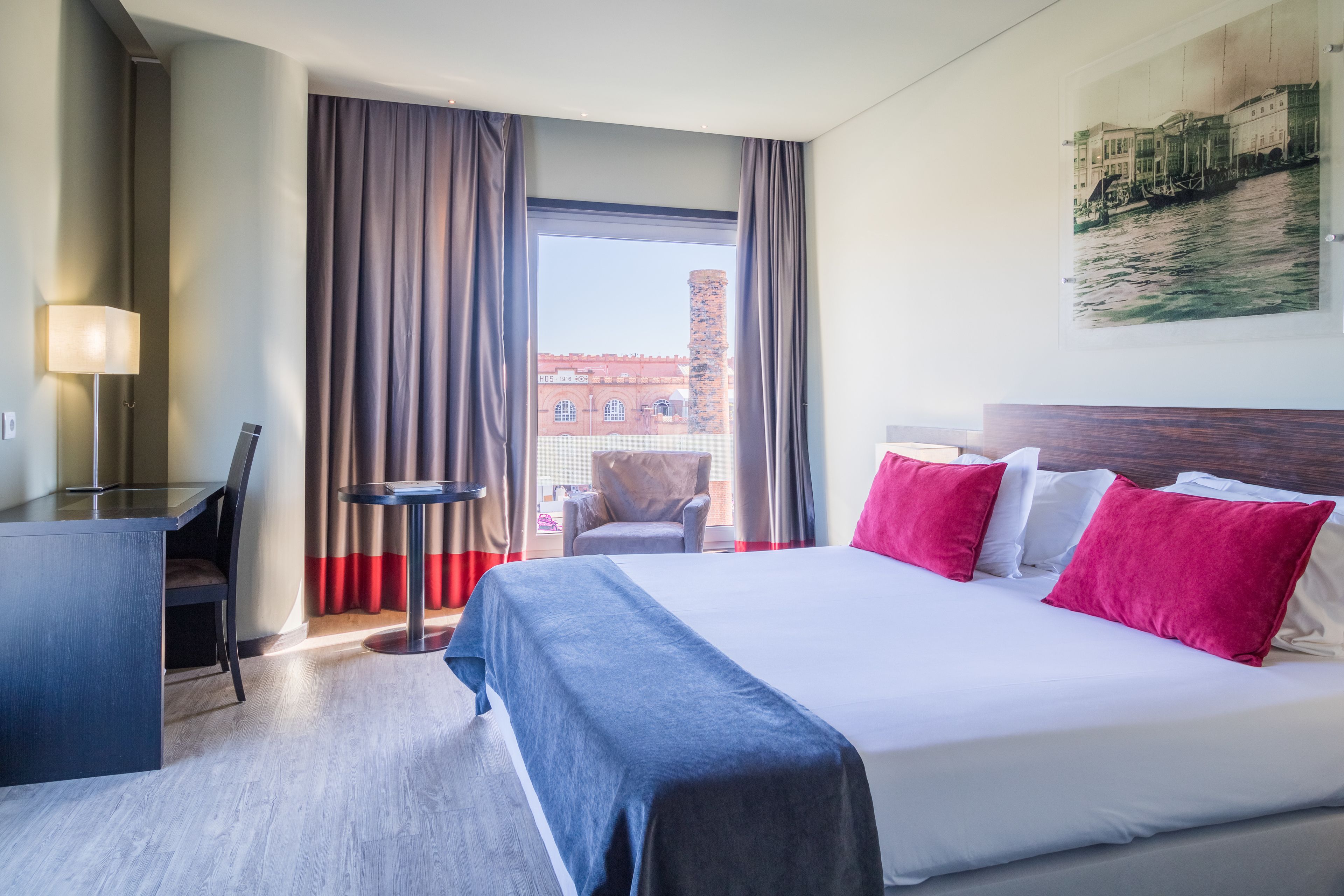 A hotel room with a bed, a desk, a chair, and a view of a building through the window.