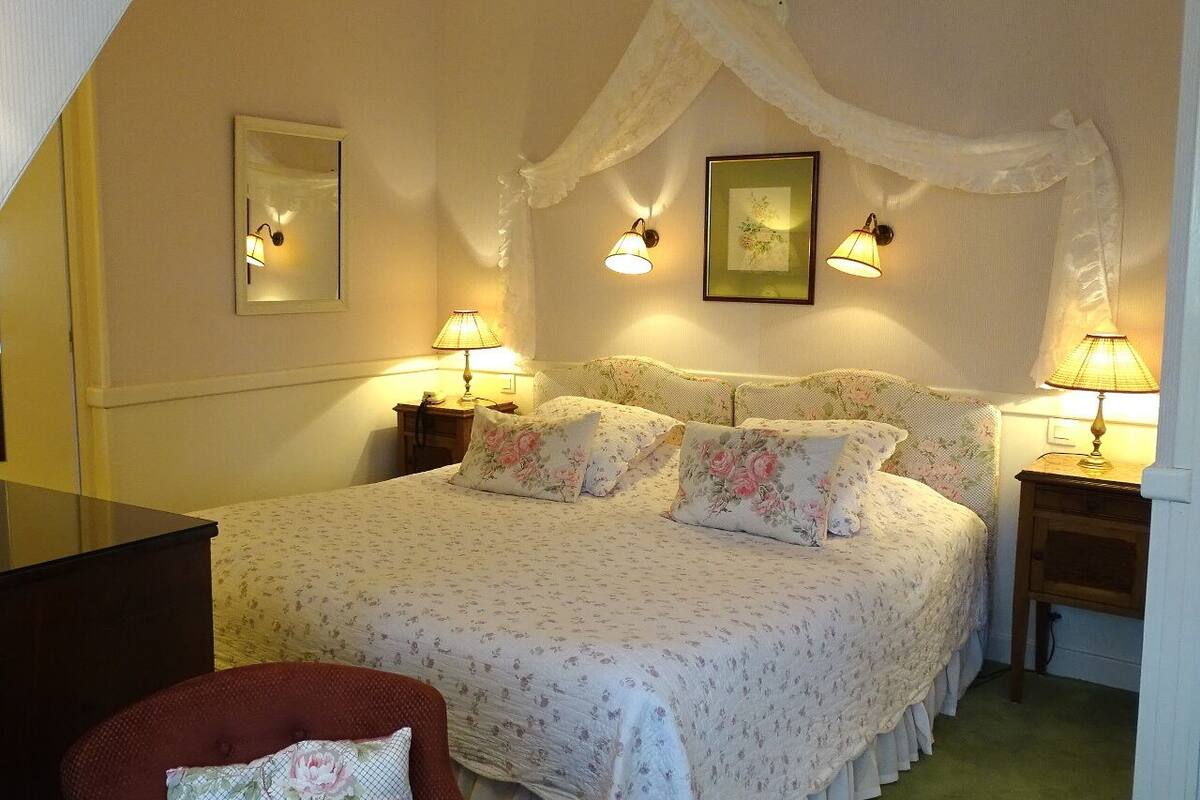 A bedroom with a canopy bed, two bedside lamps, a framed picture on the wall, and a chair with a floral pillow.