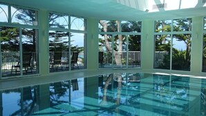 An indoor swimming pool with clear glass walls, surrounded by chairs and tables, and large windows offering views of trees and the sky. - TI AL LANNEC Hotel - Restaurant & Spa (Trébeurden)