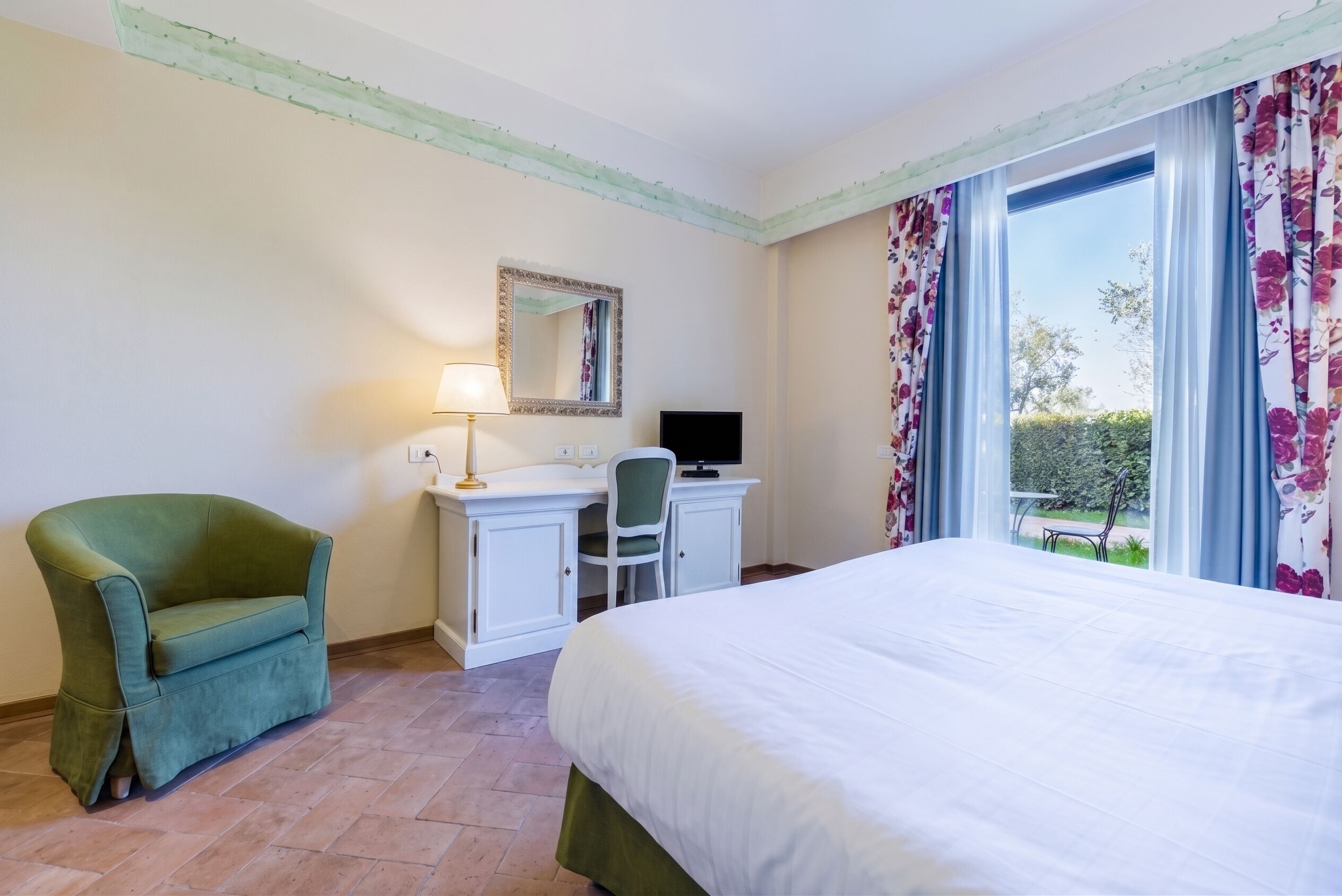 Quarto de hotel com cama, escrivaninha, computador, cadeira e janela com cortinas.