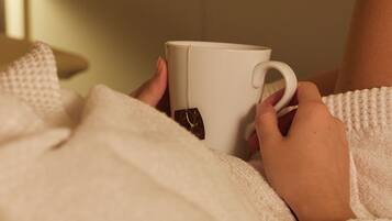 Une personne tient une tasse blanche ornée d’un motif représentant du thé, et est allongée sur un lit recouvert d’une couverture blanche.