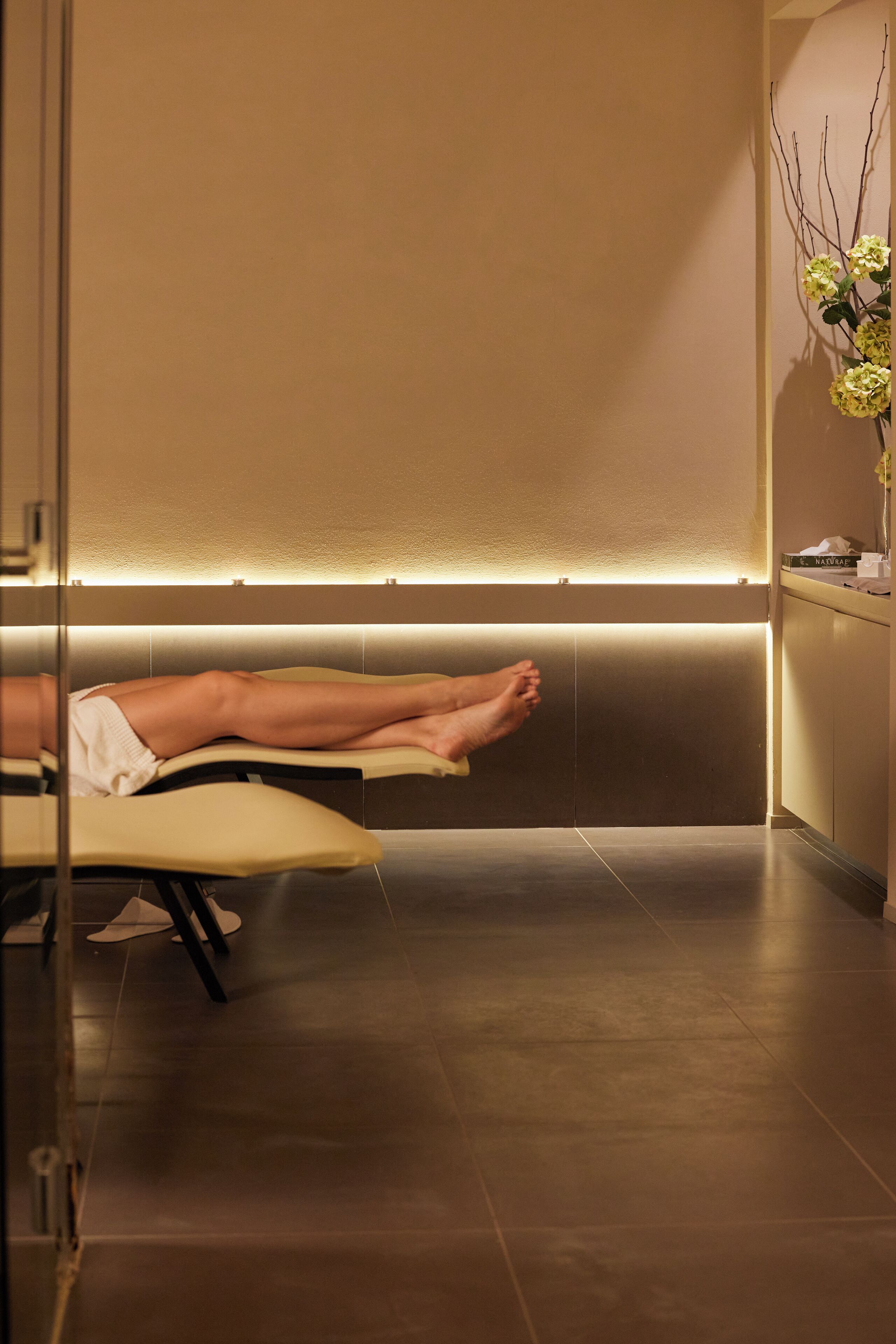 A spa room with a massage table, a glass partition, and a wall-mounted light fixture.