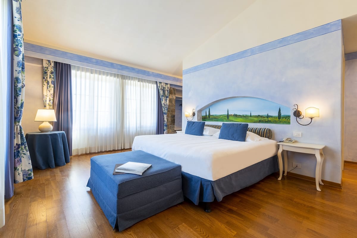 A hotel room with a large bed, a side table with a lamp, a small table with a lamp, a painting on the wall, and a view of the countryside.