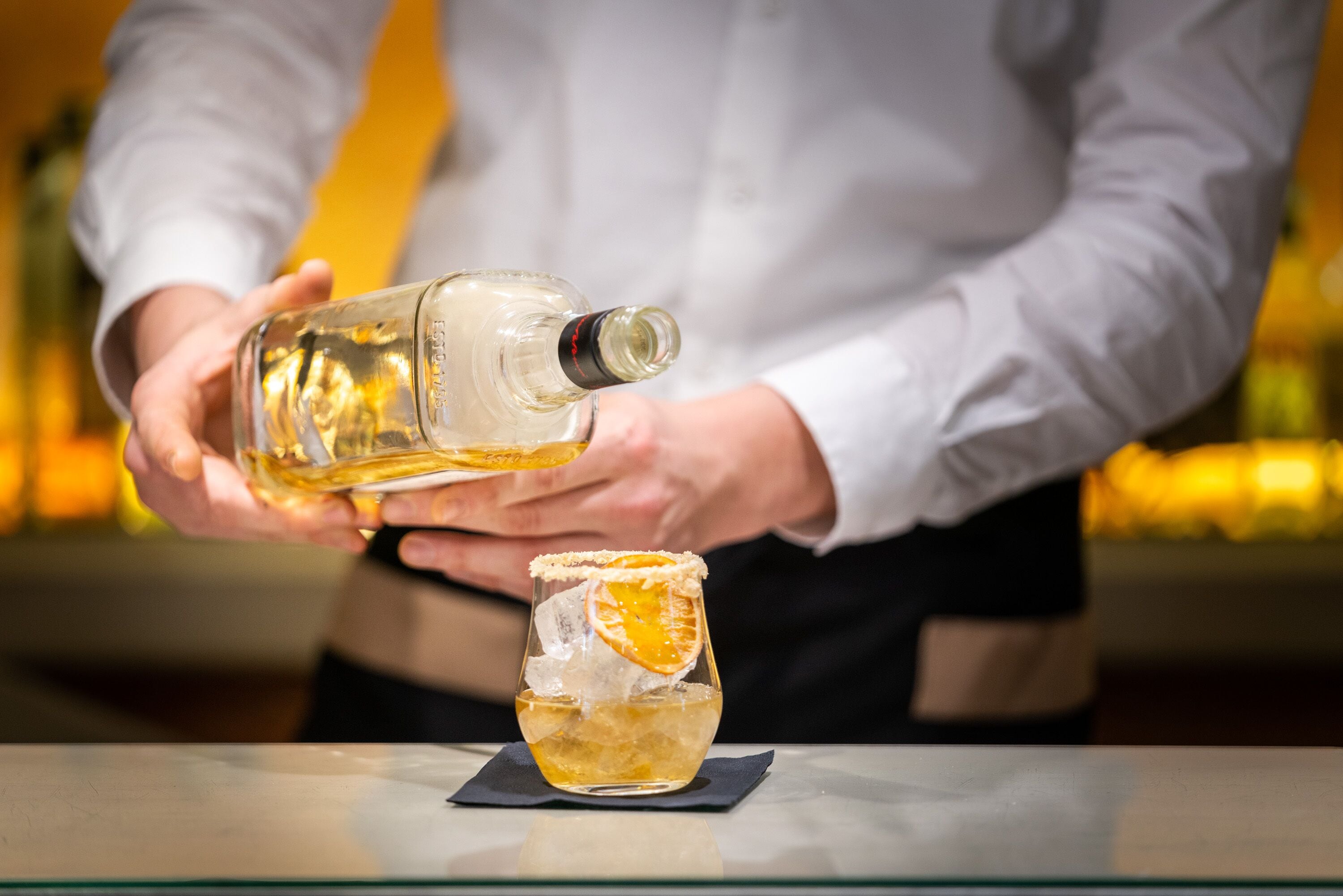 A person pouring a clear liquid into a glass with ice and an orange slice.