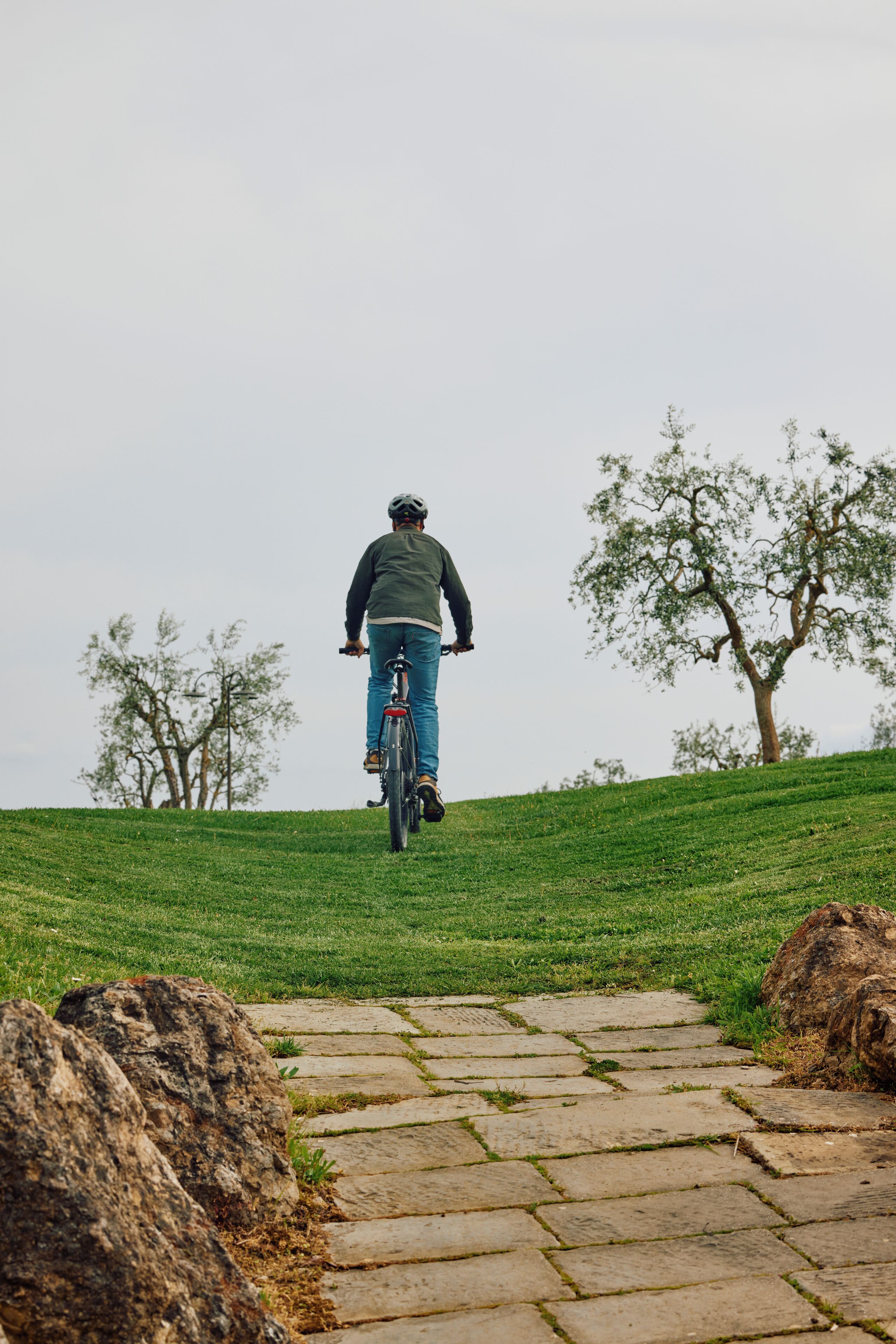 Uma pessoa anda de bicicleta por um caminho de pedra, cercada por grama e árvores.
