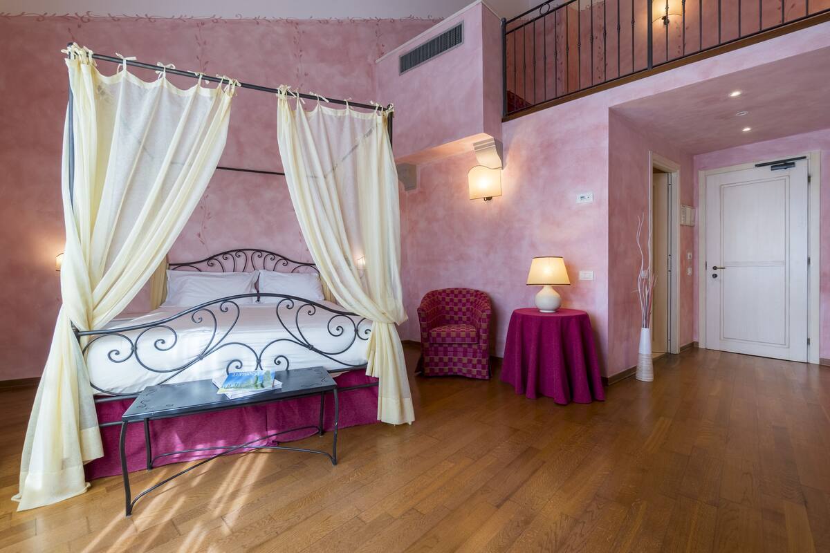 A bedroom with a four-poster bed, a small table, a chair, a lamp, and a door.