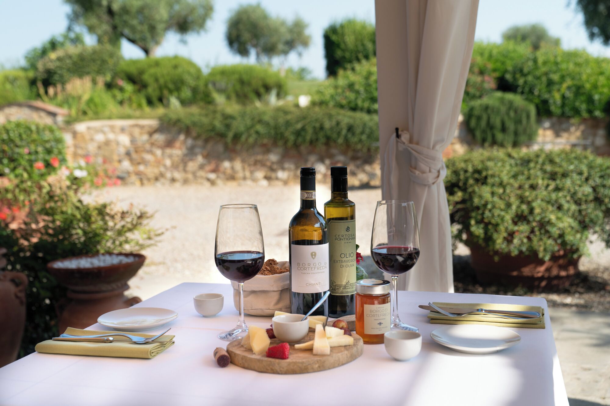 Une table dressée à l’extérieur, avec des bouteilles de vin, des verres, du fromage et des fruits.